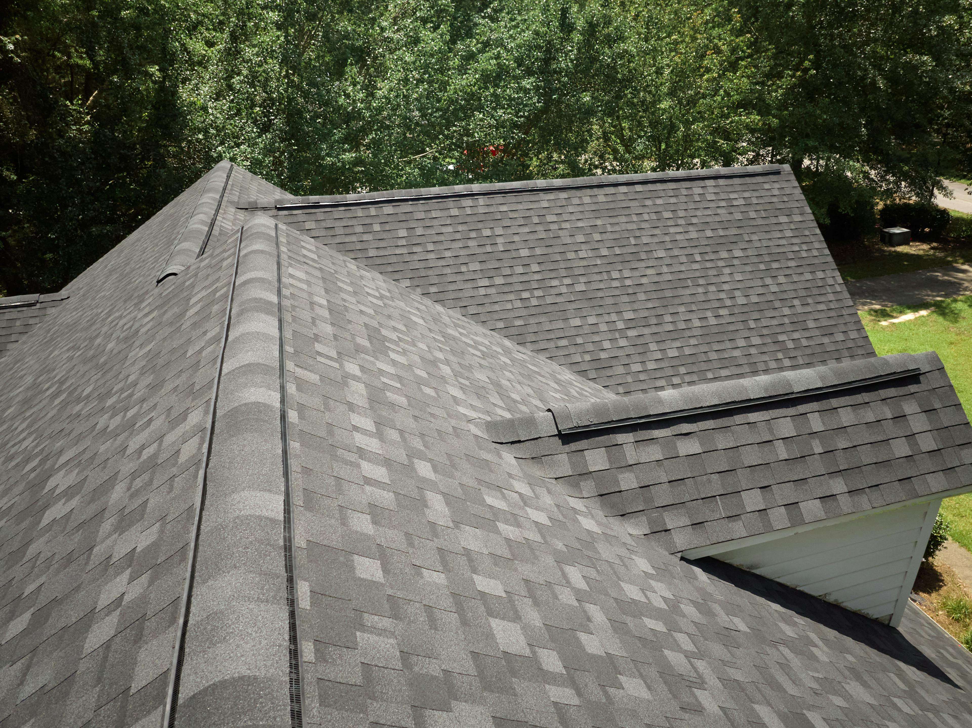 Image from atop a residential roof with granite black aphalt shingles. Green grass and trees in background.