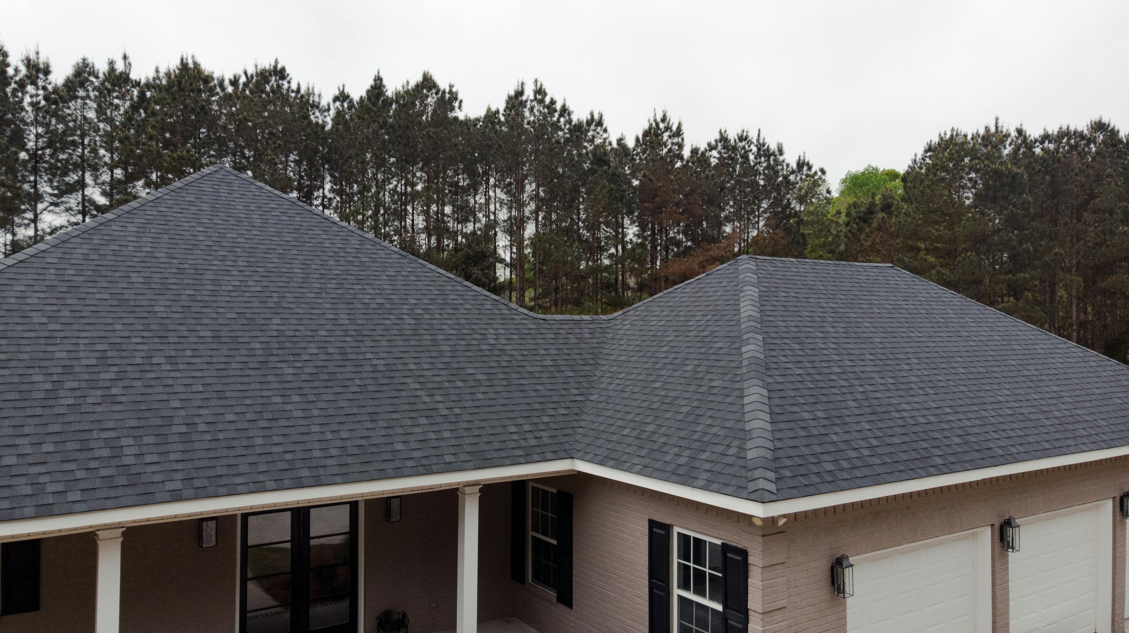 Drone image of residential roofing project. Black asphalt shingles with tree line in background.